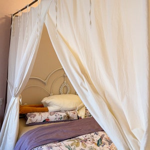 May include: A white canopy bed with a white bedspread and pillows. The bed is made up with a purple throw blanket and a floral patterned bedspread.