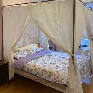 May include: A white metal canopy bed frame with a white sheer canopy and a floral patterned duvet cover. The bed is made with a white and purple pillow and a white stuffed animal.
