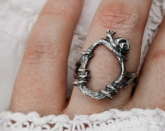 May include: A silver-toned ring featuring an open, oval-shaped design that resembles twisted branches or vines. The metal has a dark, oxidized finish in the recessed areas, highlighting the textured, organic details of the band's construction.