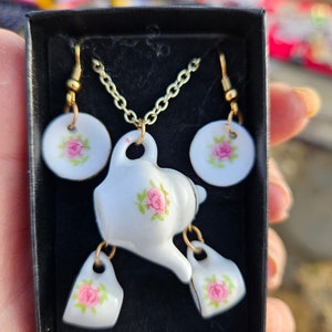May include: A white porcelain tea set necklace and earrings set with pink floral accents. The set includes a teapot pendant, two teacup charms, and two matching earrings.