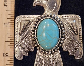 May include: A silver-toned pendant in the shape of a stylized eagle with a turquoise stone in the center. The eagle has intricate details and a geometric design.