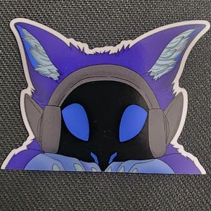 May include: A sticker featuring a stylized purple fox-like character with large blue eyes and headphones. The character has a black face and is set against a dark gray background.