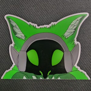 May include: A sticker featuring a stylized green fox-like character wearing gray headphones. The character has large green eyes and a black face. The sticker is on a dark gray textured surface.