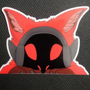 May include: A red and black sticker featuring a stylized fox-like character wearing gray headphones. The character has large red eyes and a black face. The sticker is on a black textured surface.