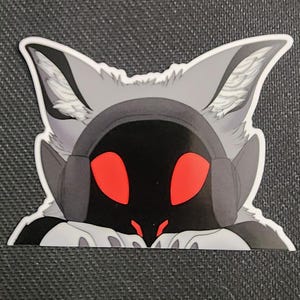 May include: A sticker featuring a stylized, gray fox-like character with red eyes and a black face. The character is wearing headphones and peeking over the bottom edge. The sticker has a white border.