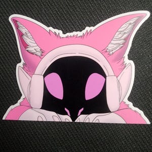 May include: A pink and white sticker featuring a stylized cat-like character with large ears and headphones. The character has a black face with pink eyes and is set against a white border.
