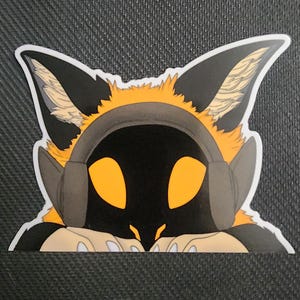 May include: A sticker featuring a stylized fox-like character with large, orange eyes and a black face wearing a headset. The character has black ears with tan fur and is set against a dark background.