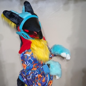 May include: A colorful Protogen partial fursuit with a black visor, blue accents, and yellow fur. The suit includes a head, hand paws, and a tail. The torso is covered in a blue patterned hoodie. The tail is black and yellow.