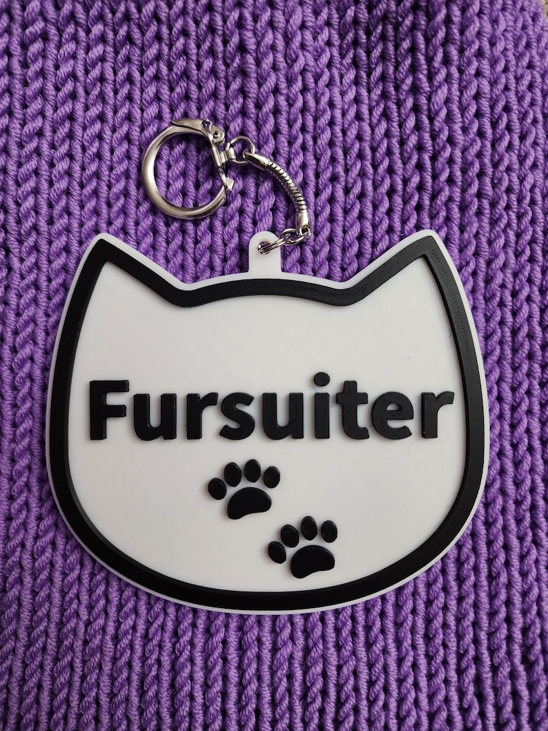 Puede incluir: Llavero blanco con forma de gato con un contorno negro y la palabra "Fursulter" en letras negras. El llavero tiene dos huellas de patas negras sobre un fondo blanco.