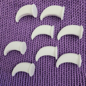 May include: Eight white plastic claw tips for knitting. The tips are curved and have a flat base. They are arranged in a semi-circle on a purple knitted surface.