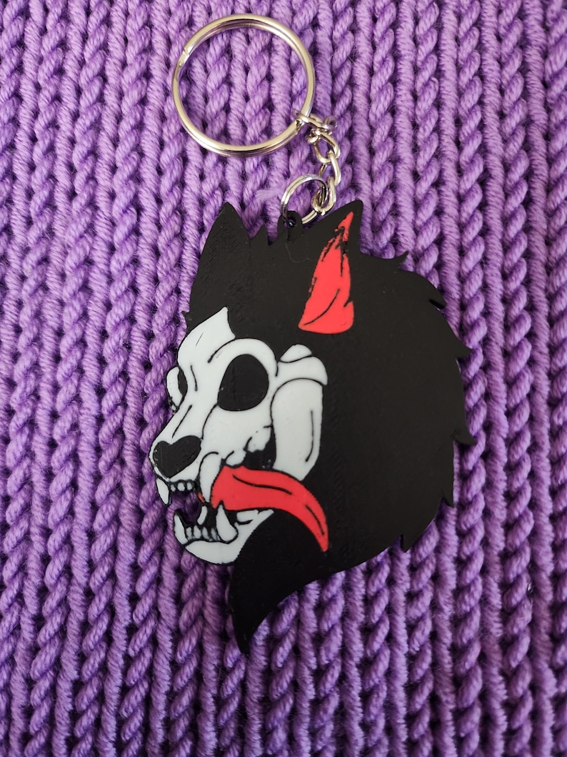 May include: Black and white wolf skull keychain with a red tongue and ear. The keychain is made of plastic and has a silver metal ring.