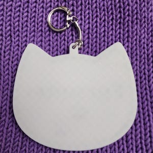 Puede incluir: Llavero blanco con forma de gato con un anillo de metal plateado. El llavero est&aacute; sobre una superficie de punto morado.