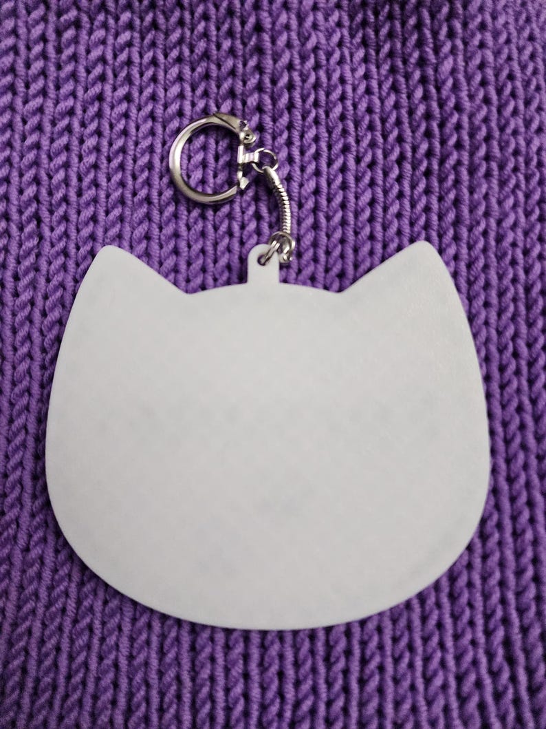 Puede incluir: Llavero blanco con forma de gato con un anillo de metal plateado. El llavero est&aacute; sobre una superficie de punto morado.