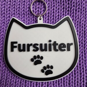 Puede incluir: Llavero blanco con forma de gato con borde negro y huellas de patas negras. El llavero tiene un anillo plateado y la palabra "Fursulter" escrita en negro.