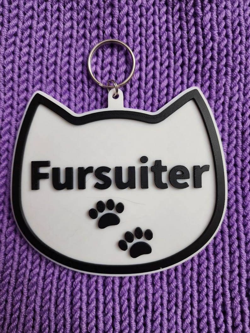 Puede incluir: Llavero blanco con forma de gato con borde negro y huellas de patas negras. El llavero tiene un anillo plateado y la palabra "Fursulter" escrita en negro.