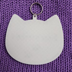 Puede incluir: Llavero blanco con forma de gato con un anillo de metal plateado. El llavero est&aacute; sobre una superficie de punto morado.