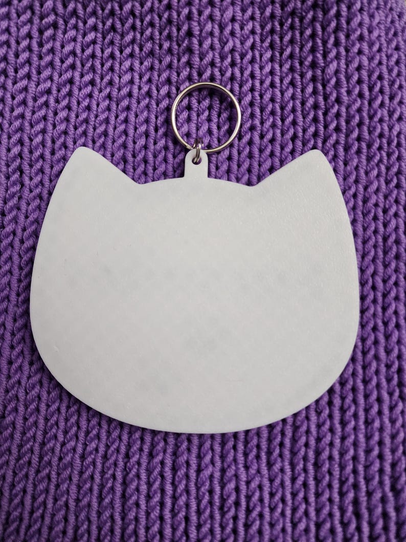 Puede incluir: Llavero blanco con forma de gato con un anillo de metal plateado. El llavero est&aacute; sobre una superficie de punto morado.