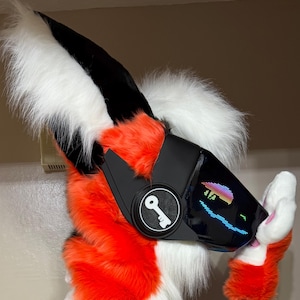 May include: A furry headpiece featuring vibrant orange and white faux fur with tall, pointed ears. The front includes a black visor with a colourful LED pixel display showing a sunset graphic and a circular side panel with a white key icon.