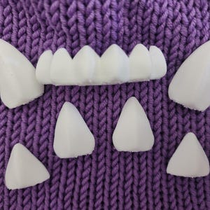 May include: A set of white plastic teeth and fangs on a purple knitted background. The teeth are arranged in a row, and the fangs are scattered around them.