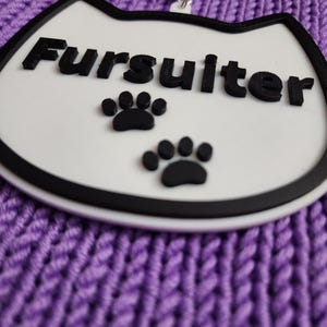 Puede incluir: Llavero blanco con forma de gato con contorno negro y la palabra "Fursulter" en letras negras. Hay dos huellas de patas negras debajo del texto.