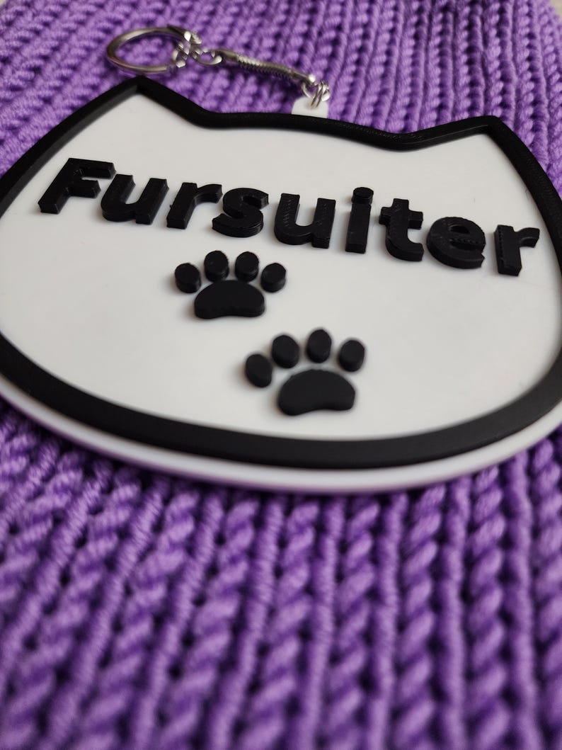 Puede incluir: Llavero blanco con forma de gato con contorno negro y la palabra "Fursulter" en letras negras. Hay dos huellas de patas negras debajo del texto.