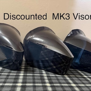 May include: Three black MK3 visors are displayed on a checkered surface. The visors have a glossy finish and a curved design. The text "Discounted MK3 Visors" is visible above the visors.
