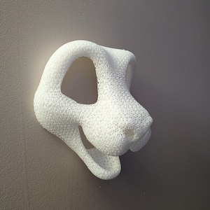 May include: A white, 3D-printed sculpture of a lion's head. The sculpture has a textured surface and an open mouth. The lion's head is mounted on a gray wall.