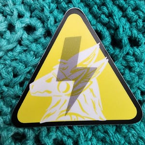 May include: Yellow warning triangle sticker with a white wolf head and a grey lightning bolt inside. The wolf's eyes are closed.