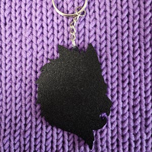 May include: Black wolf silhouette keychain with a silver metal ring and chain. The keychain is on a purple knitted surface.