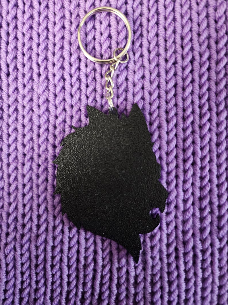 May include: Black wolf silhouette keychain with a silver metal ring and chain. The keychain is on a purple knitted surface.