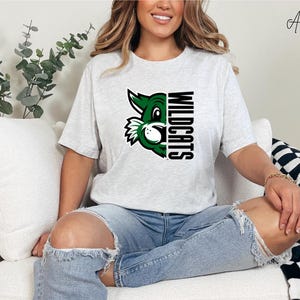 Peut inclure: T-shirt gris clair avec un motif de mascotte de chat sauvage vert et blanc et le mot "WILDCATS" en noir. Le t-shirt est de style col rond à manches courtes.