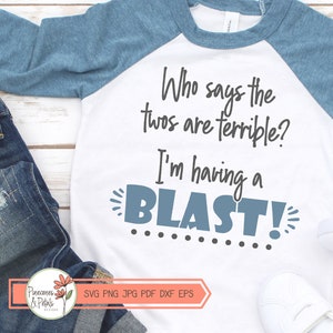 Puede incluir: Una camiseta blanca y azul de manga raglan con el texto "Who says the twos are terrible? I'm having a BLAST!" en texto negro y gris. La camiseta est&aacute; sobre una superficie de madera blanca con un par de pantalones vaqueros azules y zapatillas blancas.