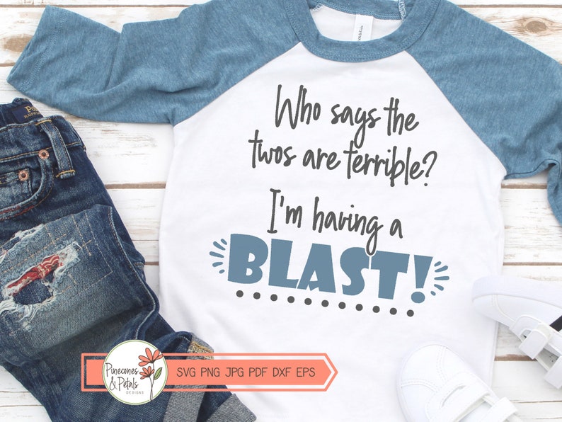 Puede incluir: Una camiseta blanca y azul de manga raglan con el texto "Who says the twos are terrible? I'm having a BLAST!" en texto negro y gris. La camiseta est&aacute; sobre una superficie de madera blanca con un par de pantalones vaqueros azules y zapatillas blancas.