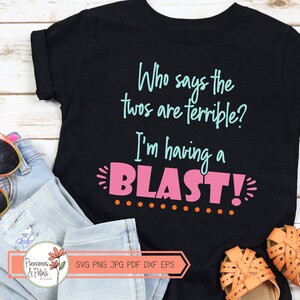 Puede incluir: Camiseta negra con el texto "Who says the twos are terrible? I'm having a BLAST!" en letras rosas y azules. La camiseta est&aacute; sobre una superficie de madera con unas gafas de sol y unos vaqueros.