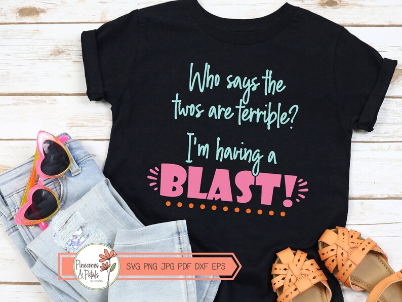 Puede incluir: Camiseta negra con el texto "Who says the twos are terrible? I'm having a BLAST!" en letras rosas y azules. La camiseta est&aacute; sobre una superficie de madera con unas gafas de sol y unos vaqueros.