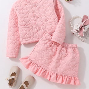 May include: A pink quilted jacket and skirt set for girls. The jacket has a round neckline, long sleeves, and button fastening. The skirt has a frilled hem and is also quilted with a heart pattern.