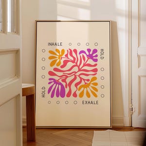 May include: A rectangular framed wall art print on a light cream background featuring a central abstract floral design in shades of pink, orange, and purple. The text INHALE, HOLD, HOLD, and EXHALE is printed in black with small circles.