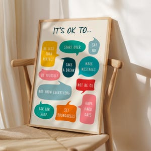 May include: A framed print with the text "IT'S OK TO..." and various phrases in colourful speech bubbles, including "Be Less Than Perfect" and "Make Mistakes." The print is displayed on a wooden chair.