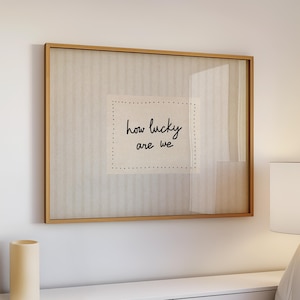 May include: A framed artwork with the text "how lucky are we" in a handwritten style. The artwork is set against a neutral background with a light wood frame. A cylindrical vase and a lamp are visible.