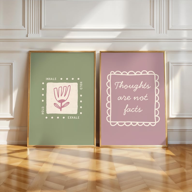 May include: Two framed art prints. One print has a sage green background with a floral design and the words "INHALE, HOLD, EXHALE". The other print has a mauve background with the words "Thoughts are not facts". Both are in gold frames.