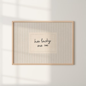 May include: A framed print with the words "how lucky are we" in a handwritten style. The text is centered on a light beige rectangle with dotted borders, set against a striped beige background, all within a light wood frame.