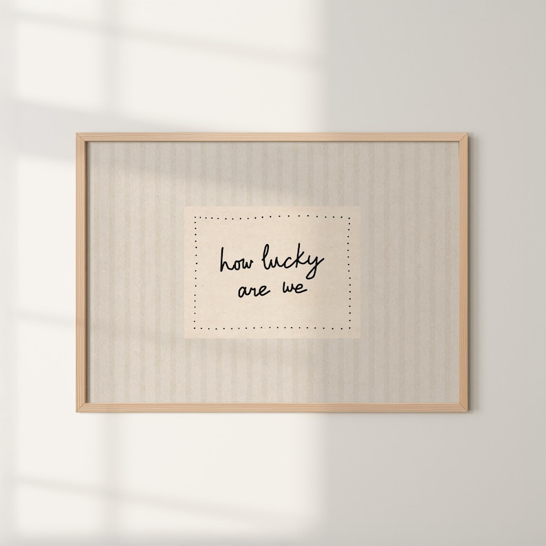 May include: A framed print with the words "how lucky are we" in a handwritten style. The text is centered on a light beige rectangle with dotted borders, set against a striped beige background, all within a light wood frame.