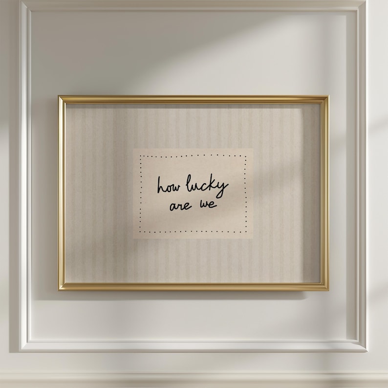 May include: A framed print with a gold-colored frame. The print features the handwritten phrase "how lucky are we" on a beige background with a dotted border. The frame is rectangular and hangs on a white wall.