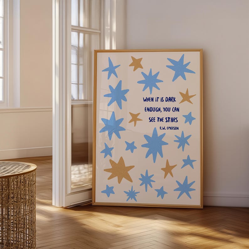 May include: A framed art print featuring a cream background with blue and gold stars. The print includes the quote "WHEN IT IS DARK ENOUGH, YOU CAN SEE THE STARS" by R.W. Emerson. The frame is a light brown color.