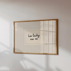May include: A framed print with the words "how lucky are we" in black script. The print is on a cream-colored background with a dotted border, set within a wooden frame. The artwork is displayed on a white wall.