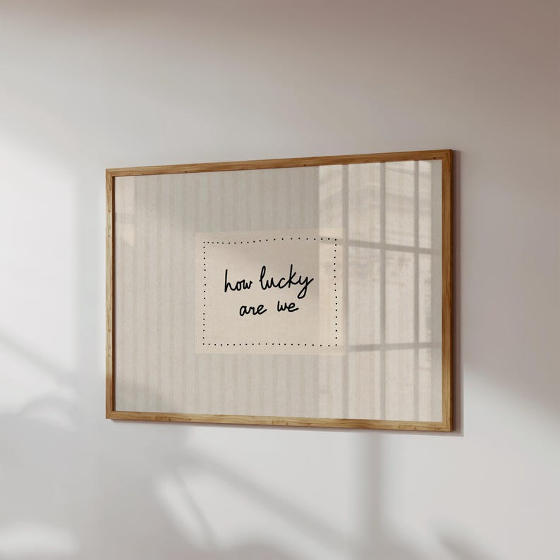 May include: A framed print with the words "how lucky are we" in black script. The print is on a cream-colored background with a dotted border, set within a wooden frame. The artwork is displayed on a white wall.