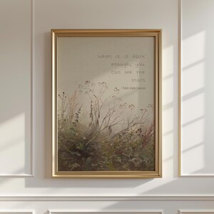 May include: A framed print with a gold-coloured frame. The artwork features a muted colour palette of greens, browns, and tans, depicting a field of plants. The text "When it is dark enough, you can see the stars" is written in a script font.