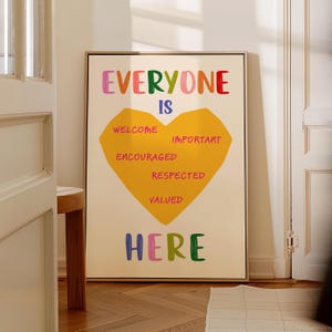 以下が含まれることがあります： カラフルな文字で「EVERYONE IS HERE」と書かれた額入りのアートプリント。中央の黄色いハート型には「WELCOME, IMPORTANT, ENCOURAGED, RESPECTED, VALUED」の文字。背景はオフホワイトです。