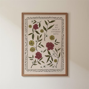 May include: A framed art print featuring a floral design with burgundy and yellow flowers, green leaves, and the text "WE ALL GROW AT DIFFERENT RATES." The print has a cream-colored background and is enclosed in a wooden frame.