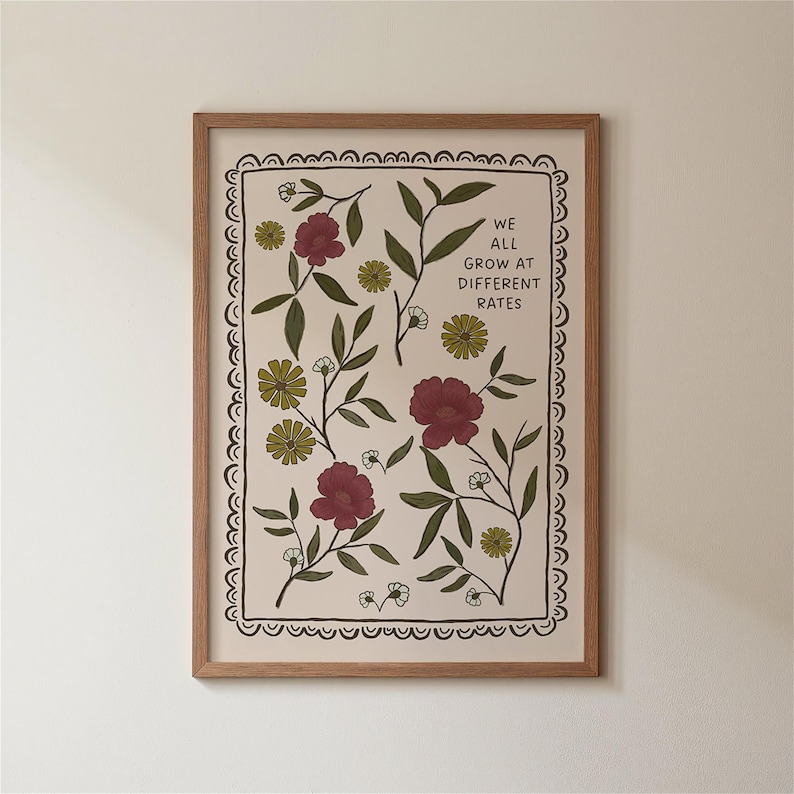 May include: A framed art print featuring a floral design with burgundy and yellow flowers, green leaves, and the text "WE ALL GROW AT DIFFERENT RATES." The print has a cream-colored background and is enclosed in a wooden frame.